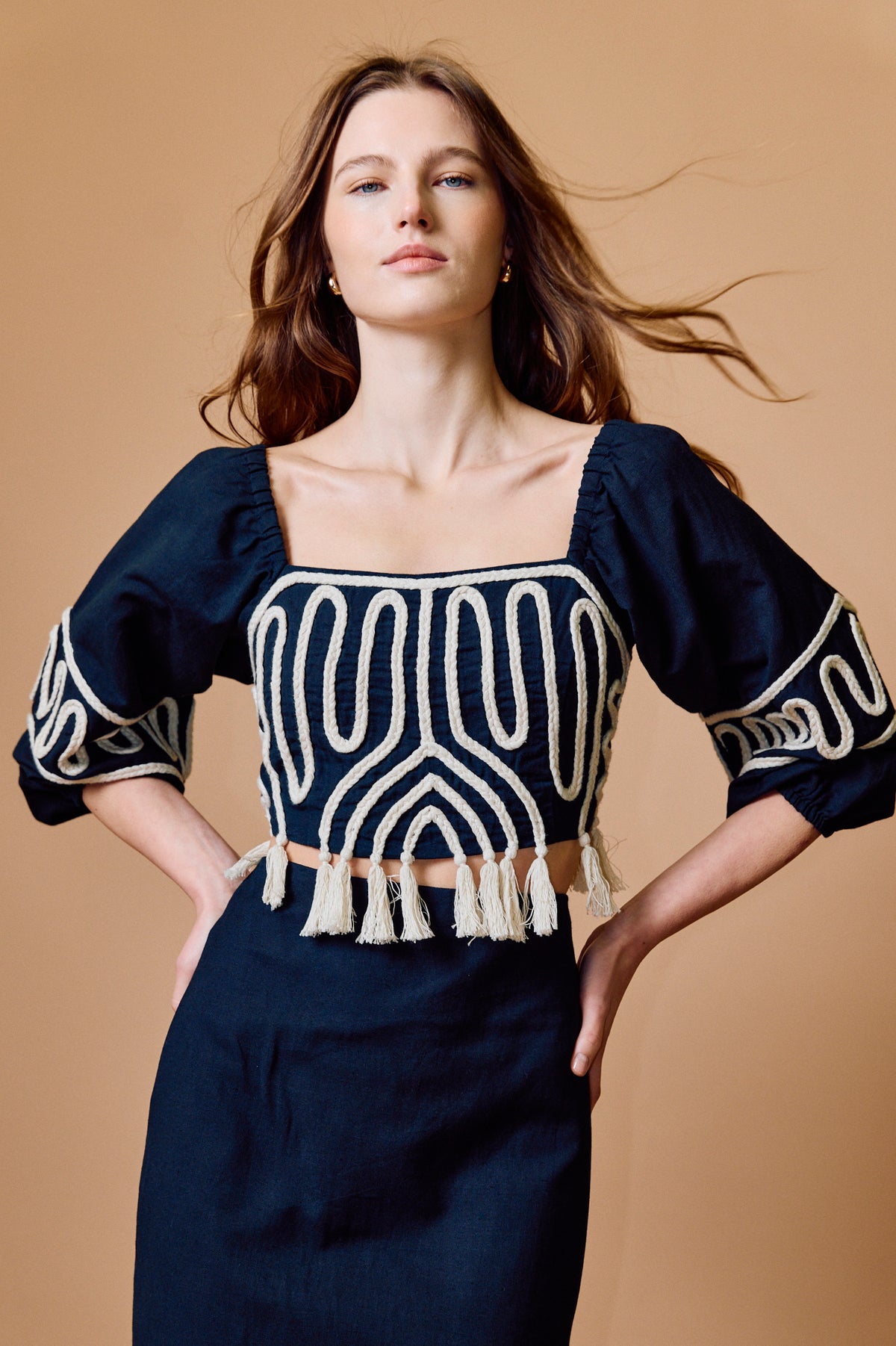 A woman modeling a black top with cream geometric embroidery, puff sleeves, and a tassel trim, paired with a matching black skirt, against a solid tan background.