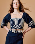 A woman modeling a black top with cream geometric embroidery, puff sleeves, and a tassel trim, paired with a matching black skirt, against a solid tan background.