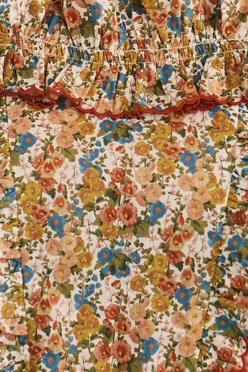 A close-up of a floral fabric with a smocked elastic waistband and ruffled trim. The pattern features a dense mix of yellow, orange, blue, and pink flowers with green leaves on a cream background.