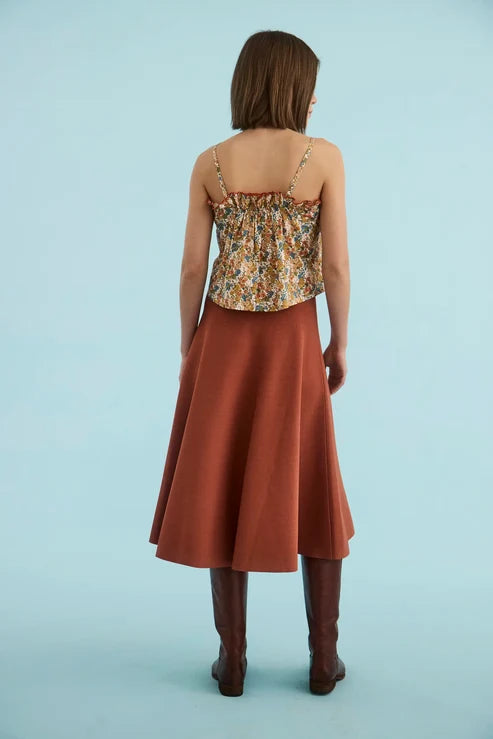 A woman shown from the back wearing a sleeveless spaghetti-strap top with a multicolor floral print and a rust-colored A-line midi skirt, paired with brown knee-high boots, against a solid light blue background.