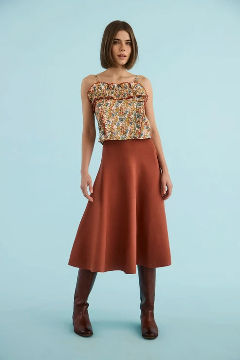 A woman modeling a sleeveless top with a multicolor floral pattern and a rust-colored satin midi skirt, paired with brown knee-high boots, against a solid light blue background.