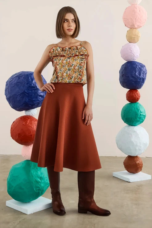 A woman stands in a studio wearing a strapless top with a multicolor floral print and a rust-brown flared skirt, paired with brown boots. She is framed by two tall stacks of colorful textured paper balls on white pedestals.