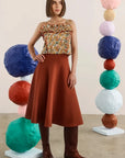 A woman stands in a studio wearing a strapless top with a multicolor floral print and a rust-brown flared skirt, paired with brown boots. She is framed by two tall stacks of colorful textured paper balls on white pedestals.