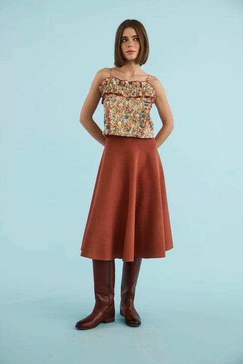 A woman stands against a light blue studio background wearing a sleeveless ruffled top with a multicolor floral print, a rust-brown A-line skirt, and dark brown knee-high boots.