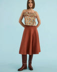 A woman stands against a light blue studio background wearing a sleeveless ruffled top with a multicolor floral print, a rust-brown A-line skirt, and dark brown knee-high boots.