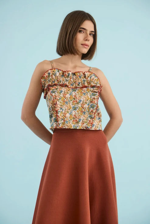 A woman modeling a sleeveless top with a multicolor floral print and ruffled trim, paired with a rust-colored skirt, against a solid light blue background.