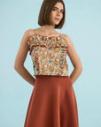 A woman modeling a sleeveless top with a multicolor floral print and ruffled trim, paired with a rust-colored skirt, against a solid light blue background.