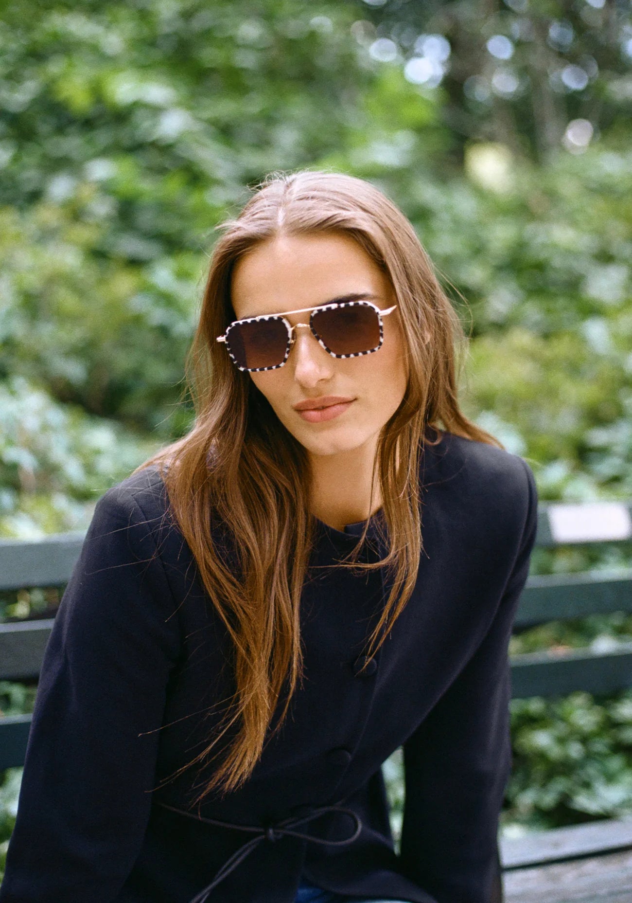 Woman wearing Emery 18K + Matte Domino Sunglasses sitting on a bench with greenery in the background