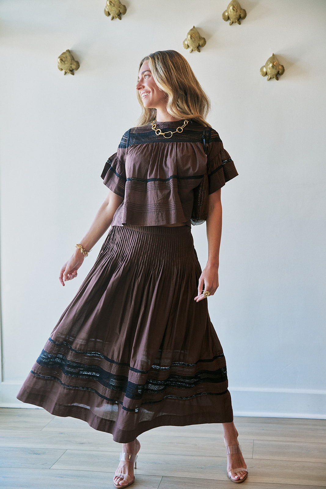 A woman modeling a brown two-piece outfit consisting of a short-sleeve ruffled top and a matching pleated maxi skirt with dark blue horizontal stripes, standing against a white wall with gold decorative accents.
