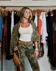A woman models a matching camouflage jacket and pants set with a white tank top and a brown belt, standing in front of a clothing rack.
