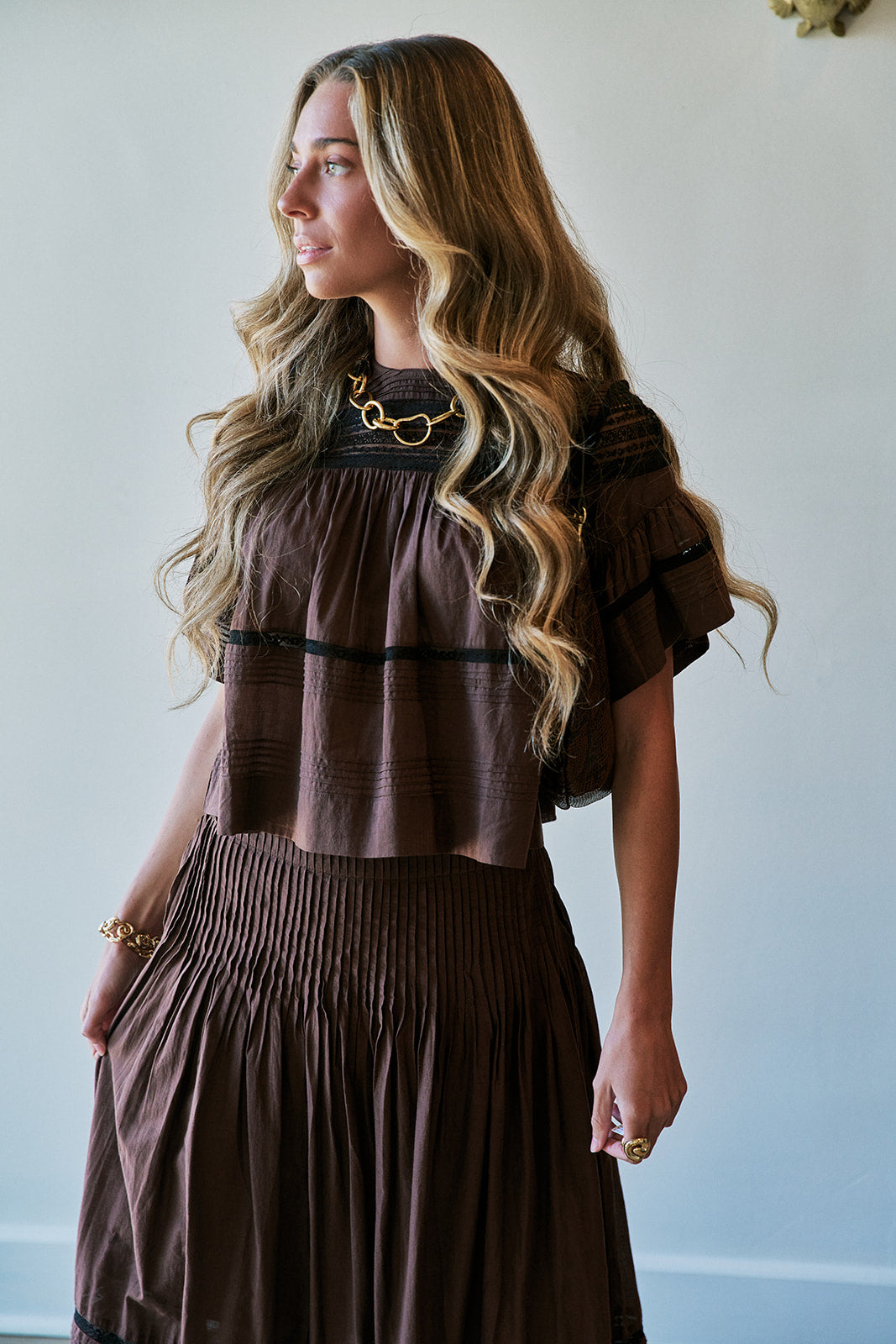 A woman with long wavy blonde hair wearing a brown tiered top and matching pleated skirt, styled with gold jewelry against a plain light gray wall.