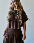 A woman with long wavy blonde hair wearing a brown tiered top and matching pleated skirt, styled with gold jewelry against a plain light gray wall.