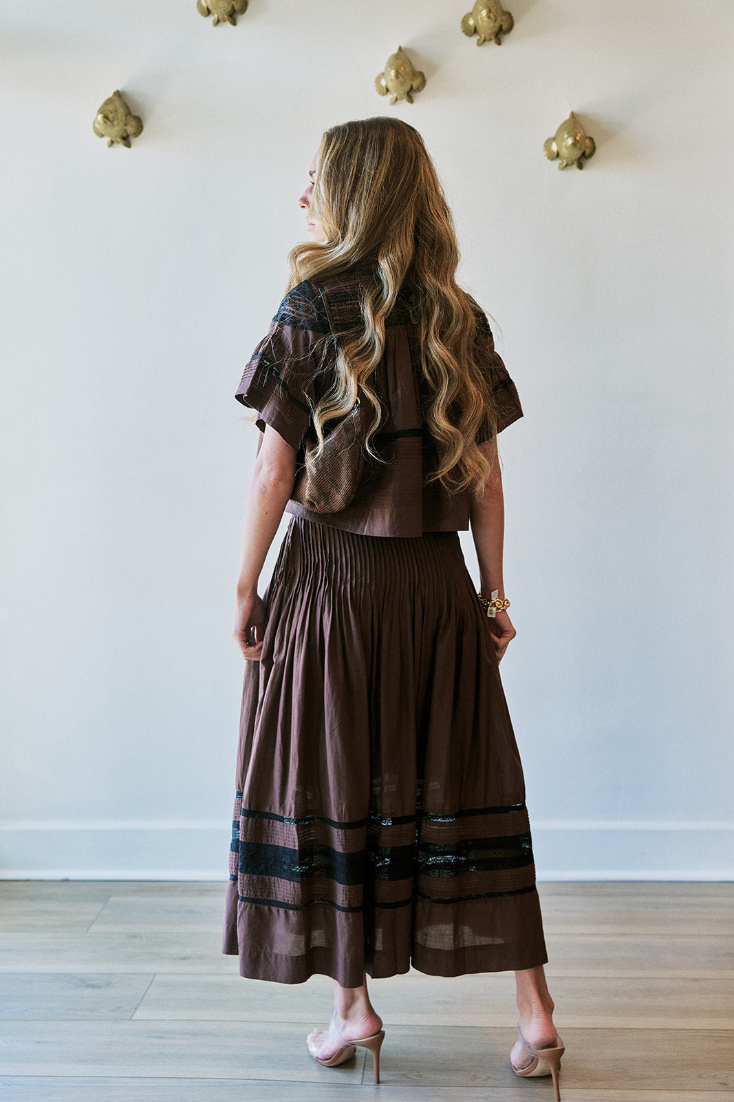 A woman with long wavy blonde hair is shown from behind wearing a brown two-piece outfit consisting of a short-sleeve cropped top and a matching pleated maxi skirt with black horizontal stripes near the hem. She is standing in a room with a light-colored wall decorated with gold wall art and light wood flooring.