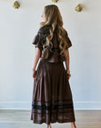 A woman with long wavy blonde hair is shown from behind wearing a brown two-piece outfit consisting of a short-sleeve cropped top and a matching pleated maxi skirt with black horizontal stripes near the hem. She is standing in a room with a light-colored wall decorated with gold wall art and light wood flooring.