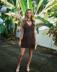 A woman stands outdoors on a paved path surrounded by large tropical green leaves and a white fence, wearing a sleeveless dark brown mini dress with decorative stitching and holding a small metallic clutch.