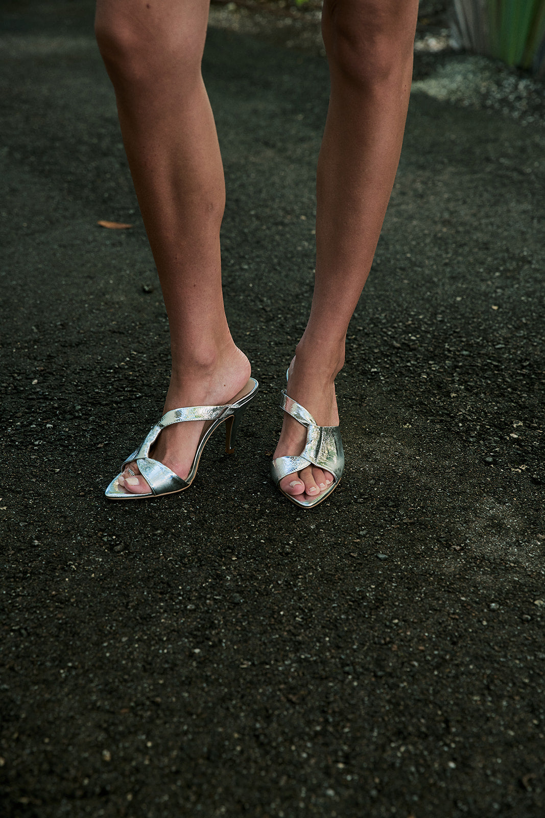A close-up of a person's legs wearing metallic silver strappy slingback high-heeled sandals on a dark asphalt surface.