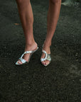 A close-up of a person's legs wearing metallic silver strappy slingback high-heeled sandals on a dark asphalt surface.