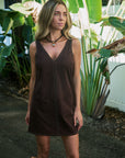 A woman wearing a sleeveless brown mini dress with studded trim, standing outdoors in front of large tropical green leaves.