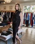 A woman modeling a sleeveless black feather-trimmed maxi dress with a thigh-high slit, standing inside a clothing boutique with racks of garments in the background.