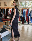 A woman modeling a sleeveless black feather-trimmed maxi dress with a thigh-high slit, standing inside a clothing boutique with racks of garments in the background.