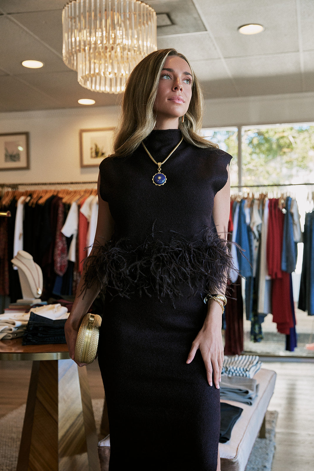 A woman models a black sleeveless mock-neck dress with a feathered waistband in a boutique setting. She is wearing a gold pendant necklace with a blue stone and holding a gold clutch.