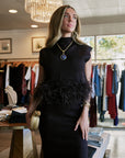 A woman models a black sleeveless mock-neck dress with a feathered waistband in a boutique setting. She is wearing a gold pendant necklace with a blue stone and holding a gold clutch.