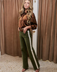 A woman modeling a brown satin long-sleeve button-up shirt and olive green wide-leg pants with a cream stripe down the side, standing in front of brown curtains.