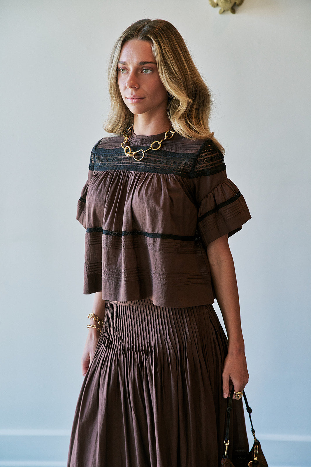 A woman wearing a brown tiered ruffle top and matching pleated skirt, styled with a chunky gold chain necklace and holding a small handbag, set against a plain light gray wall.