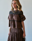 A woman wearing a brown tiered ruffle top and matching pleated skirt, styled with a chunky gold chain necklace and holding a small handbag, set against a plain light gray wall.