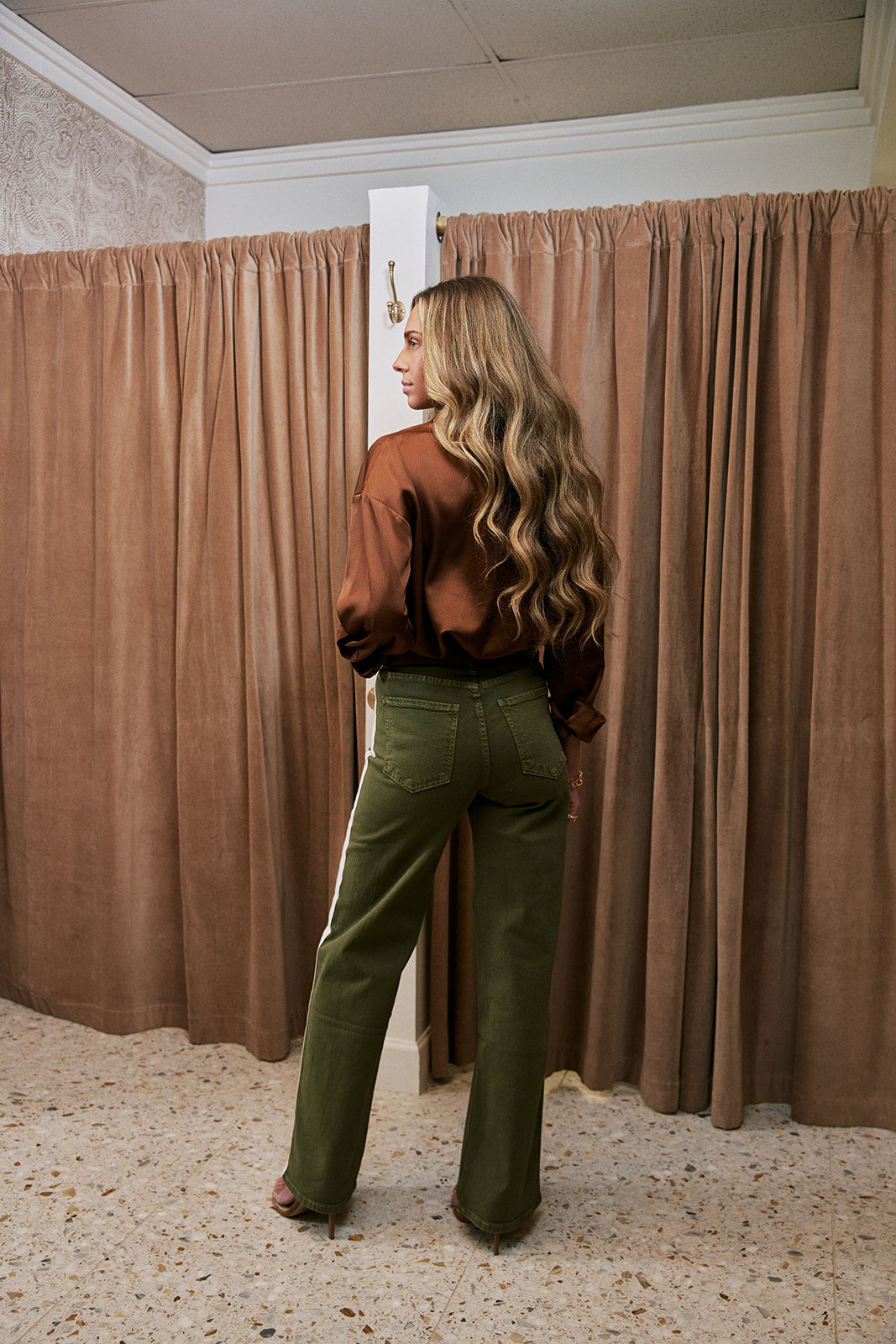 A woman with long wavy blonde hair stands in a fitting room wearing olive green wide-leg pants with a white side stripe and a brown satin blouse, viewed from behind as she looks over her outfit. The background features brown curtains and a speckled floor.