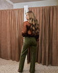 A woman with long wavy blonde hair stands in a fitting room wearing olive green wide-leg pants with a white side stripe and a brown satin blouse, viewed from behind as she looks over her outfit. The background features brown curtains and a speckled floor.