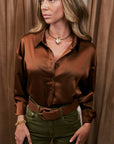 A woman wearing a brown satin button-down shirt and olive green pants, standing in front of brown curtains.
