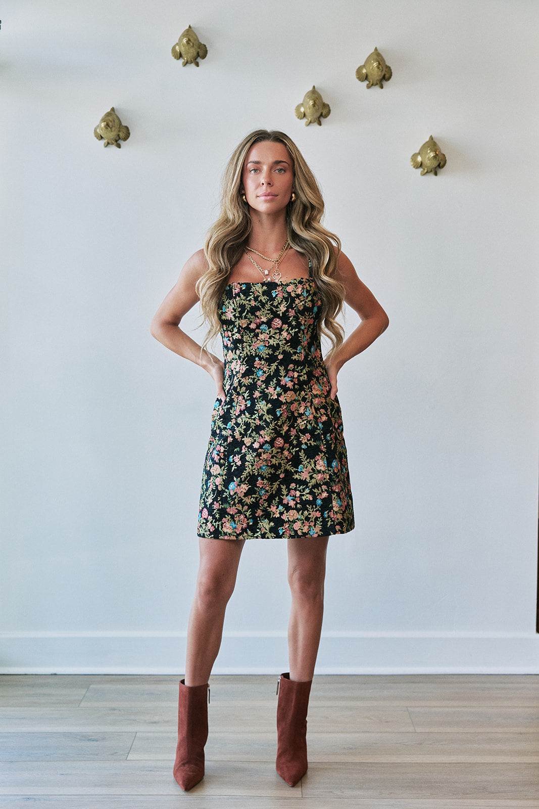 A woman stands indoors wearing a strapless black floral mini dress with a dense multicolor floral print and brown pointed-toe ankle boots. The background is a white wall with decorative gold wall hooks and light wood flooring.