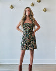 A woman stands indoors wearing a strapless black floral mini dress with a dense multicolor floral print and brown pointed-toe ankle boots. The background is a white wall with decorative gold wall hooks and light wood flooring.