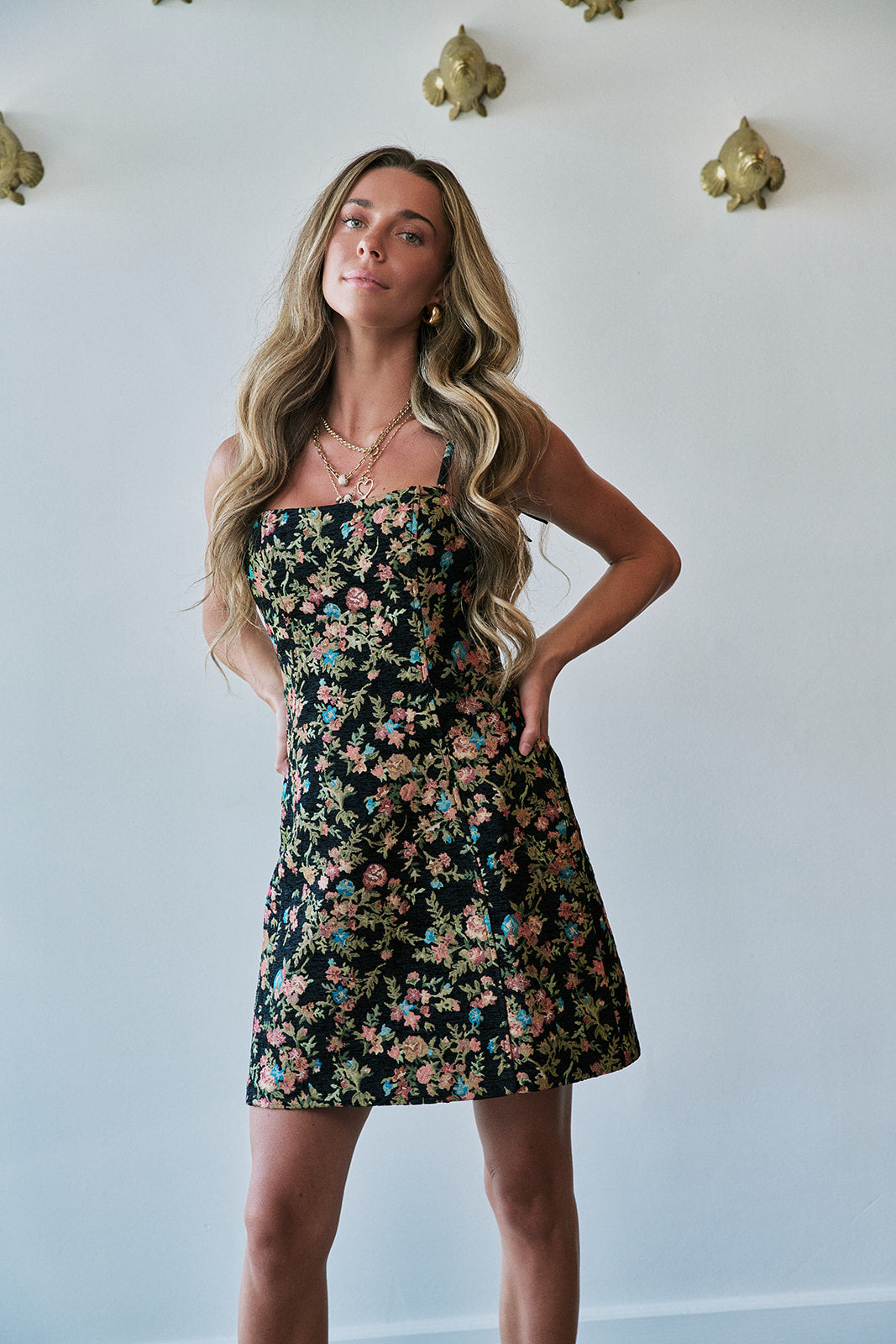 A woman modeling a strapless black mini dress with a colorful floral print, standing against a white wall with gold wall decor.