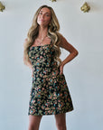 A woman modeling a strapless black mini dress with a colorful floral print, standing against a white wall with gold wall decor.