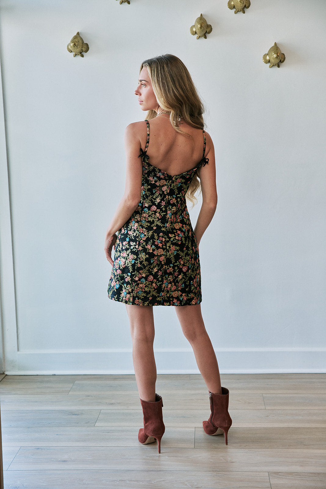 A woman stands with her back to the camera wearing a short black floral mini dress with thin straps and brown heeled ankle boots, posed in a room with a white wall and light wood floor.