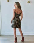 A woman stands with her back to the camera wearing a short black floral mini dress with thin straps and brown heeled ankle boots, posed in a room with a white wall and light wood floor.