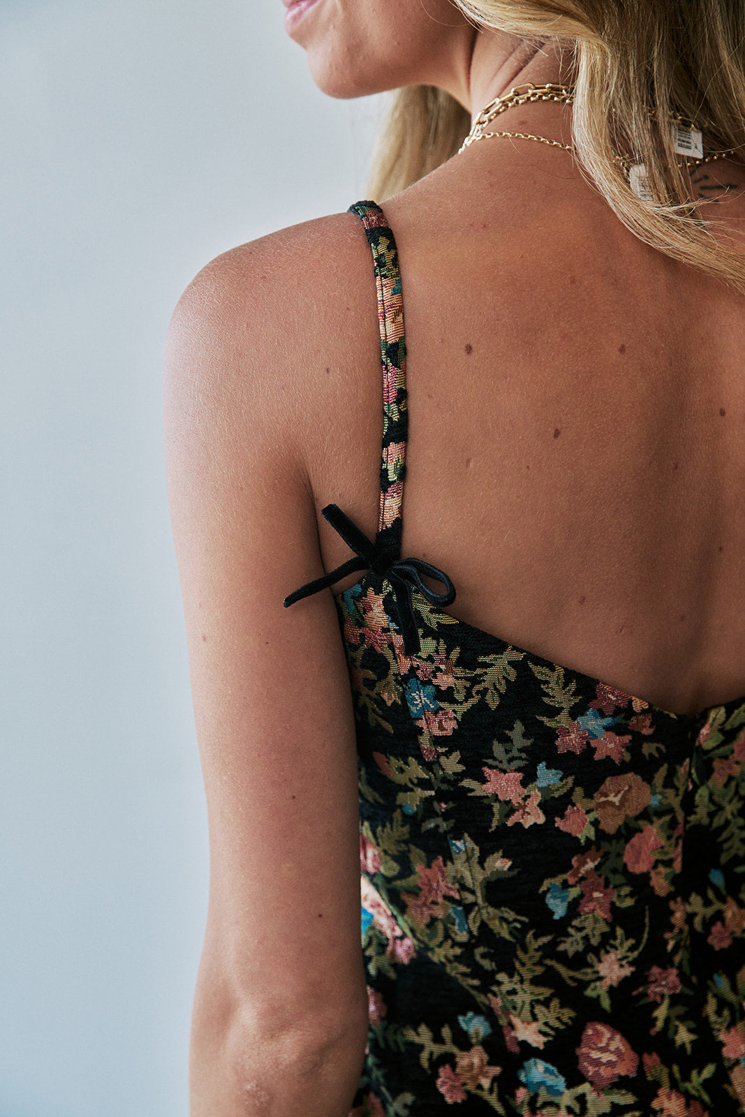 Close-up back view of a woman wearing a floral spaghetti-strap dress with black ribbon ties at the shoulders, against a light gray background.