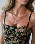 A close-up of a woman wearing layered gold necklaces, including a heart-shaped charm and a small animal charm, styled with a black floral spaghetti-strap dress.