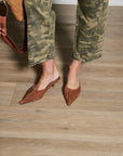 A person wearing brown suede pointed-toe mule flats and olive green camouflage pants, standing on a light wood plank floor.