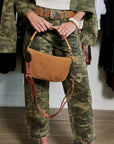A person wearing a camouflage jacket and matching pants holds a tan suede shoulder bag with a brown leather strap in a boutique setting.