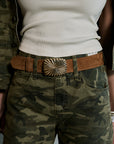 A close-up of a person wearing a brown suede belt with a large metallic sunburst buckle, paired with camouflage pants and a white ribbed top.