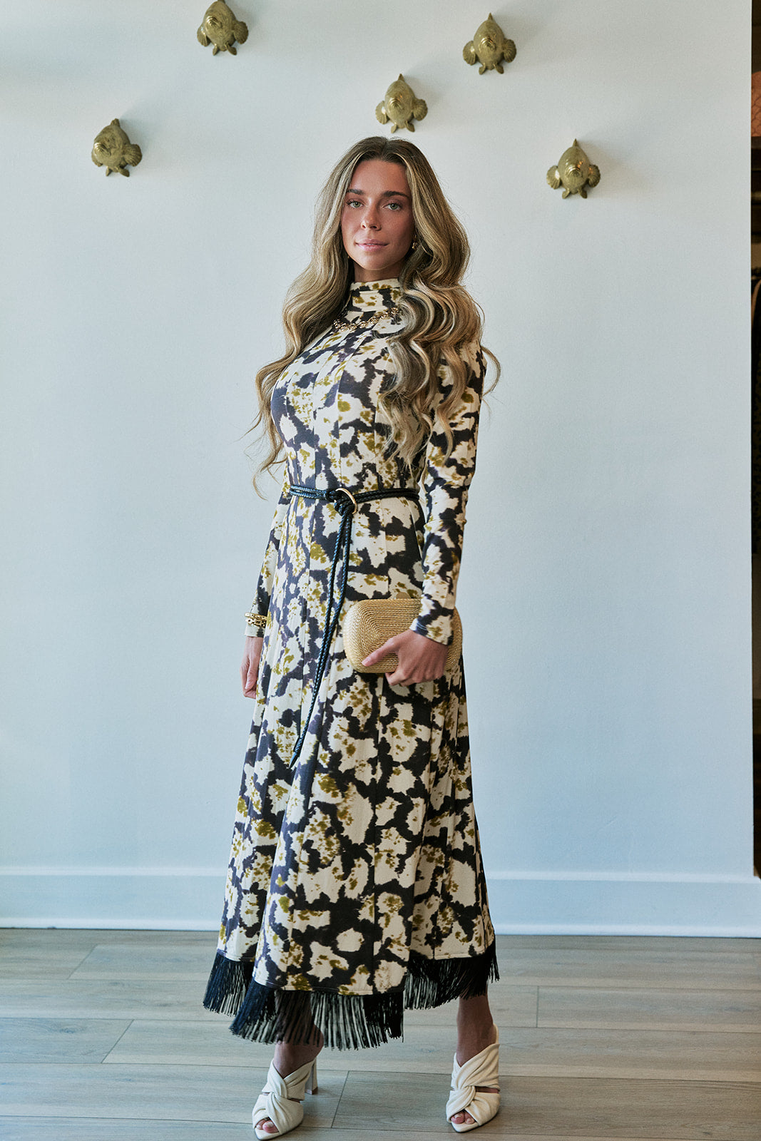 A woman modeling a long-sleeve black midi dress with a beige and white floral pattern, a high ruffled collar, a matching waist tie, and fringe detailing along the hem. She is holding a small beige clutch and wearing white heeled sandals, standing against a white wall with gold decorative wall pieces.
