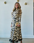 A woman modeling a long-sleeve black midi dress with a beige and white floral pattern, a high ruffled collar, a matching waist tie, and fringe detailing along the hem. She is holding a small beige clutch and wearing white heeled sandals, standing against a white wall with gold decorative wall pieces.