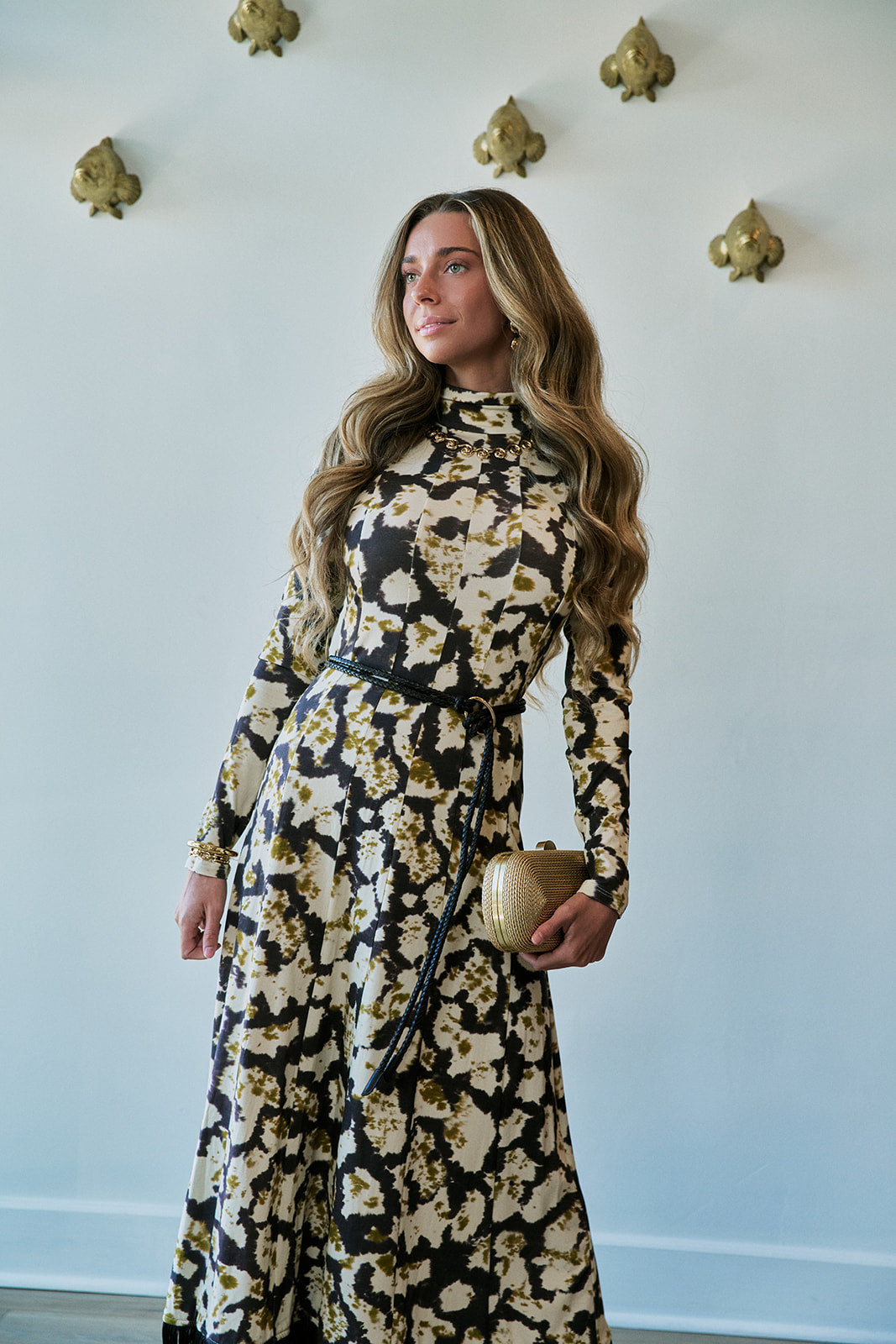 A woman modeling a long-sleeve high-neck maxi dress with a black and beige floral pattern, holding a small gold clutch against a light blue wall with gold wall decor.