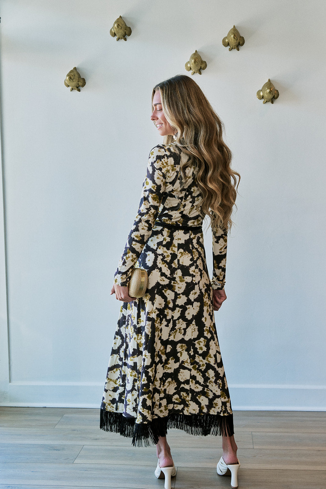 A woman wearing a long-sleeve black midi dress with a beige and gold floral pattern and fringe hem, holding a small gold clutch, standing in a room with a white wall and light wood floor.