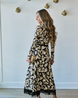 A woman wearing a long-sleeve black midi dress with a beige and gold floral pattern and fringe hem, holding a small gold clutch, standing in a room with a white wall and light wood floor.