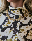 A close-up of a model wearing a gold spiral chain necklace and matching gold hoop earrings, paired with a high-neck blouse featuring a black, olive green, and cream tie-dye pattern.
