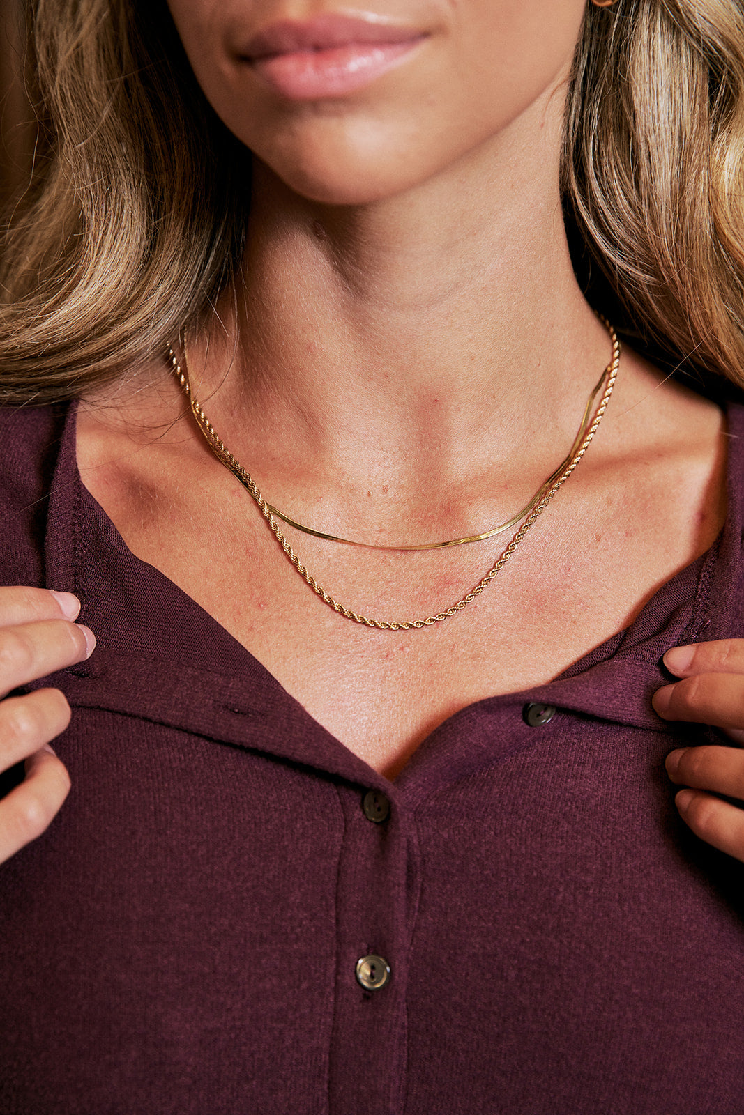 A close-up of a woman wearing a double-layered gold necklace over a dark plum button-up shirt.